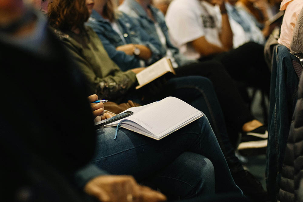 People are listening to a speaker. The photo shows their knees and most have an open Bible or notebook on their lap. Some have a phone and are taking notes.